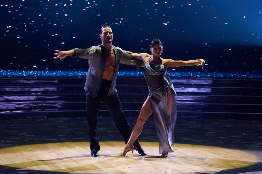 Val Chmerkovskiy and Jenna Dewan mirror each other with their inside arms crossed and their outside arms extended, wrists flipped. They roll their hips back to extend their inside legs to the side, Dewans' crossed over Chmerkovskiy's.