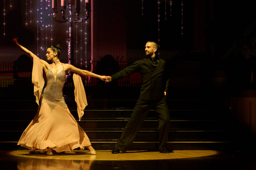Maura Higgins and Mark Ballas dance onstage in a dramatic spotlight, holding hands while swaying away from each other. Higgins extends her other arm up, her face turned away.