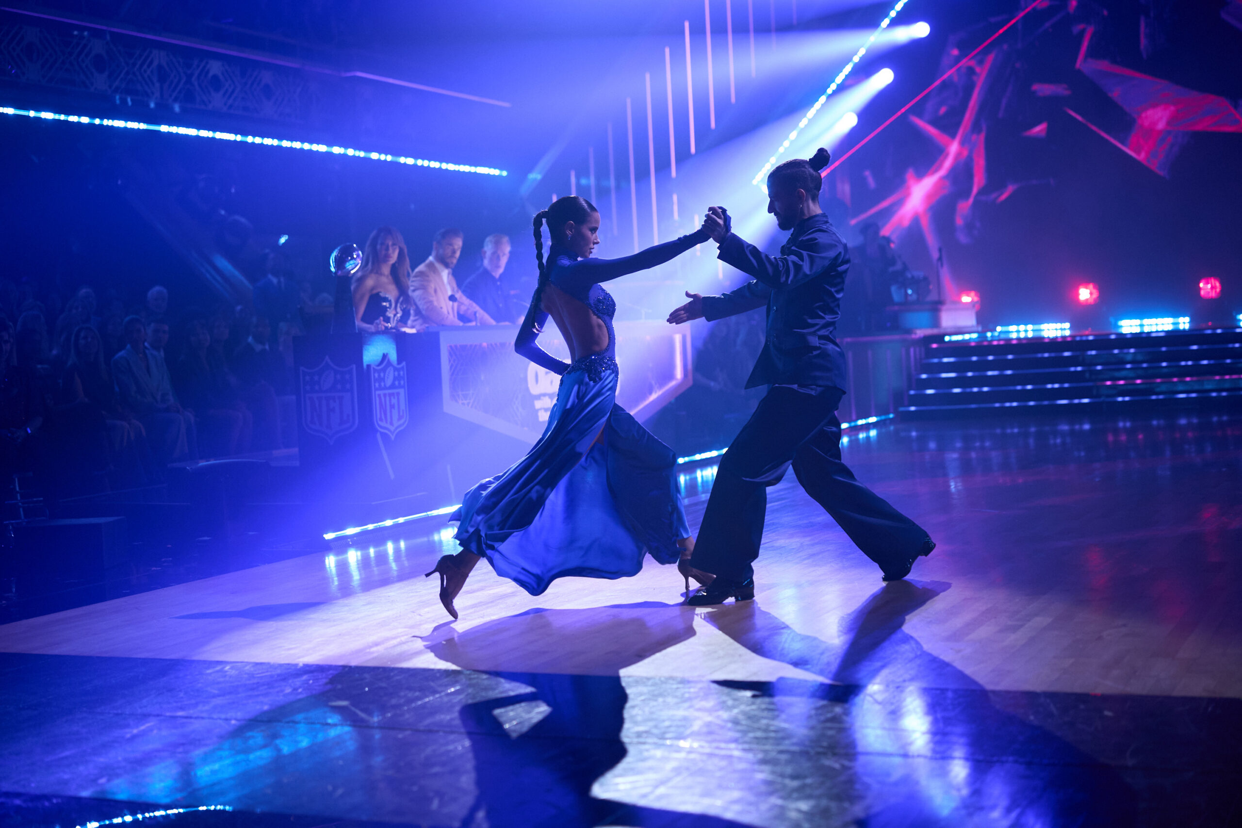 In dark blue lighting, Maura Higgins steps forward as Mark Ballas steps back; they face each other and walk into a tango frame with long, deep strides.