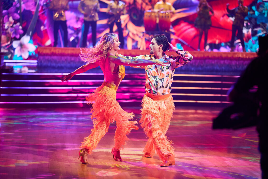 Rylee Arnold and Connor Wood mirror each other, crossing one leg over with a twist in the upper body. Wood bends one arm up so his hand touches the back of his head. They wear matching orange fringe pants.