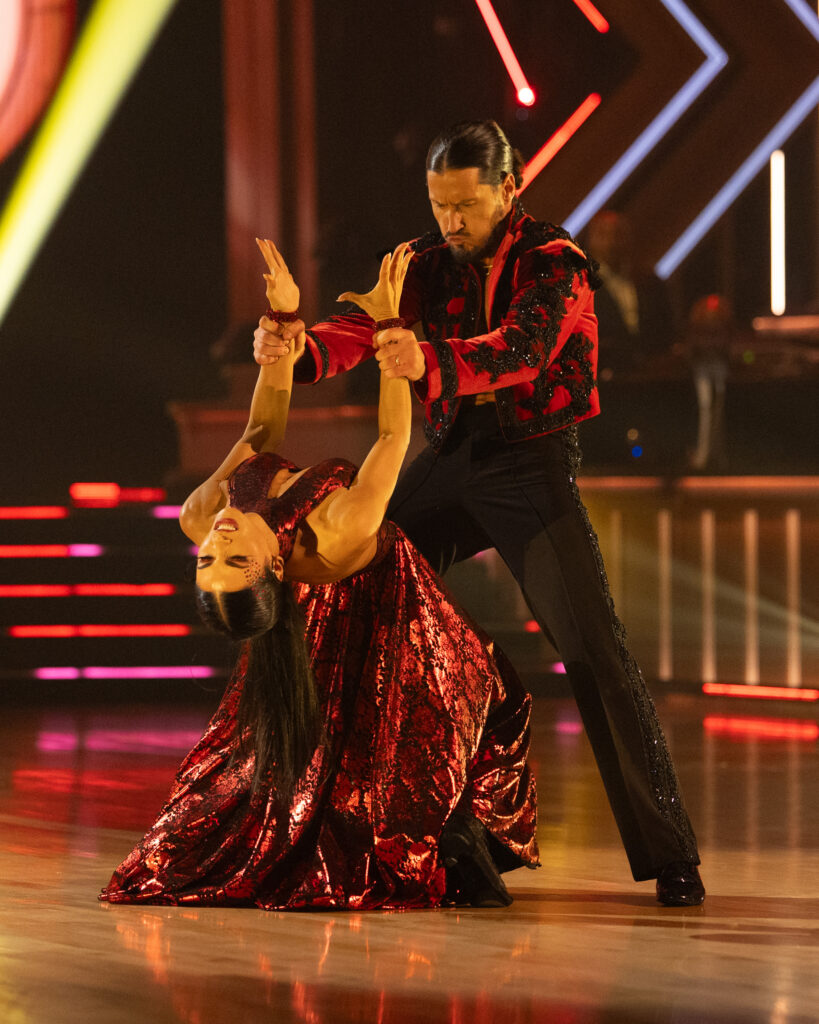 Val Chmerkovskiy stands wide-legged and holds Jenna Dewan's wrists as she does a deep hinge backward, head thrown back and arms held strong toward the ceiling.