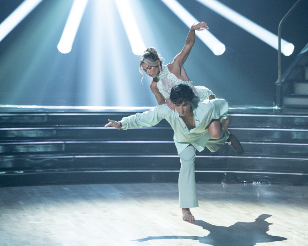 Ezra Frech stands in arabesque, leaning forward to support Daniella Karagach, who leans across his back with one arm raised.