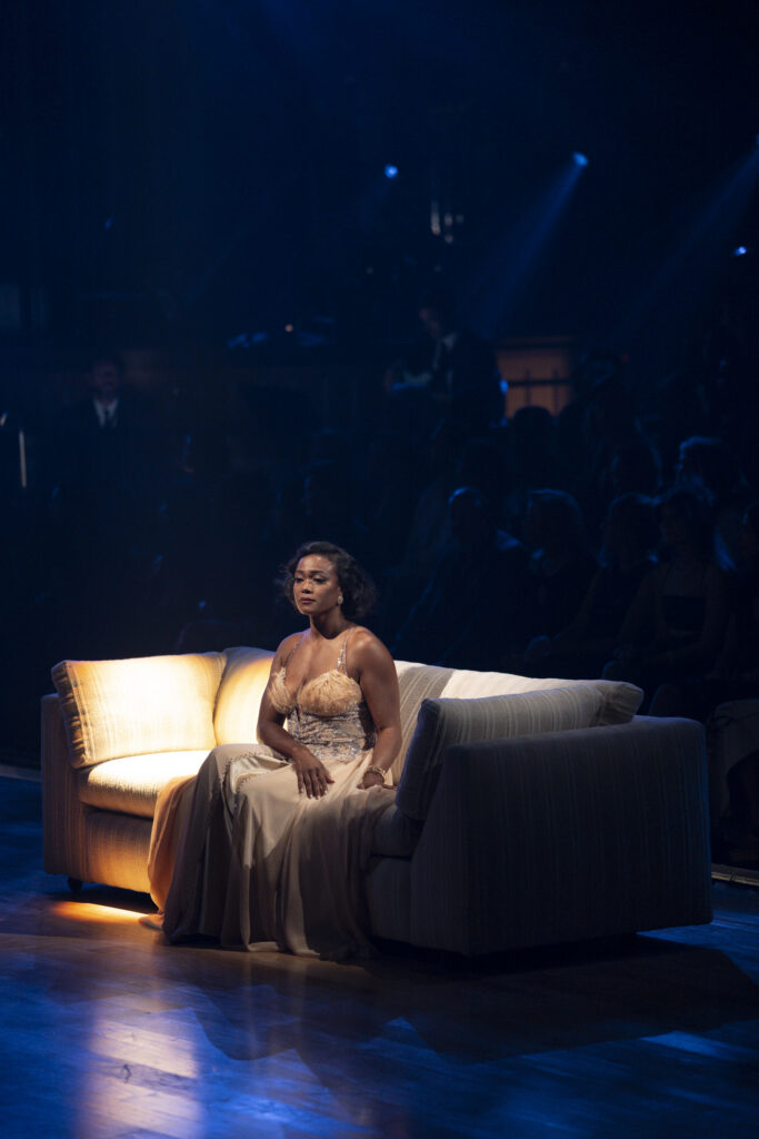 Tatyana Ali, wearing a light tan evening gown, sits on a couch with a spotlight illuminating an empty seat next to her. She looks wistfully off into the distance.