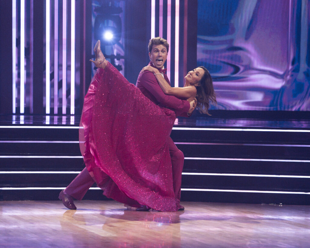 Jackson Olson holds his mother around her waist as he dips her to one side. She lifts her front leg and wraps an arm around her son's shoulder. They wear sparkling magenta costumes.