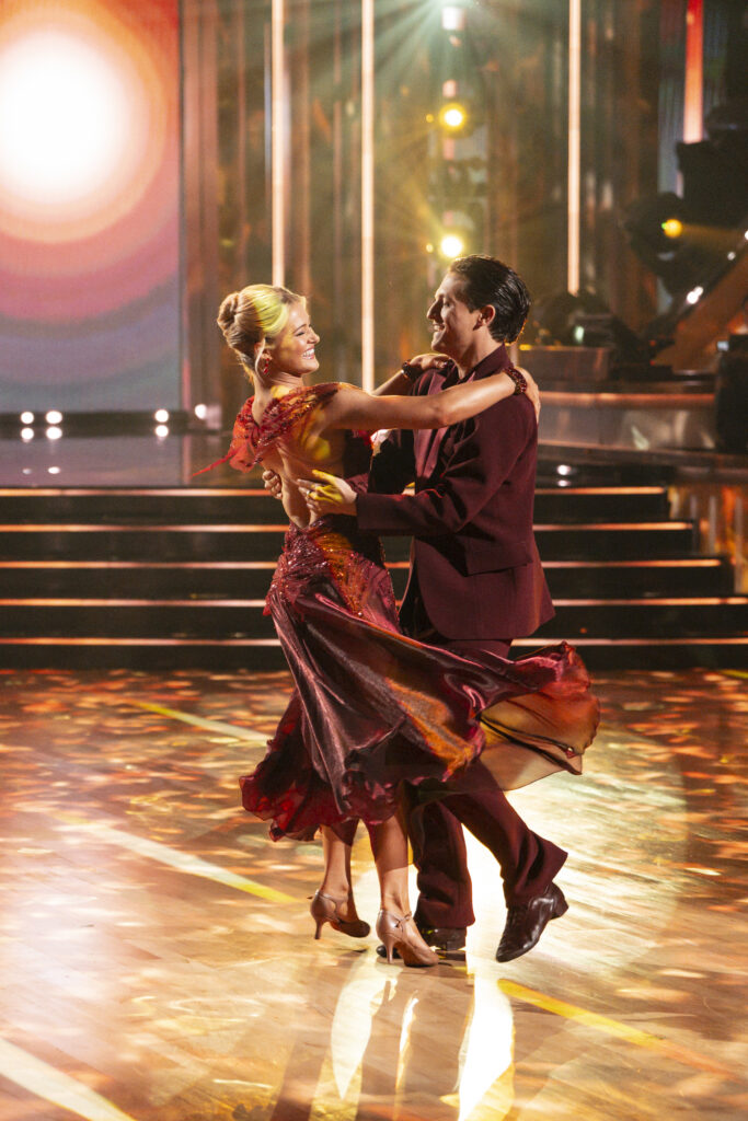 Rylee Arnold and Connor Wood smile at each other in frame as they dance a Viennese Waltz in maroon evening wear.