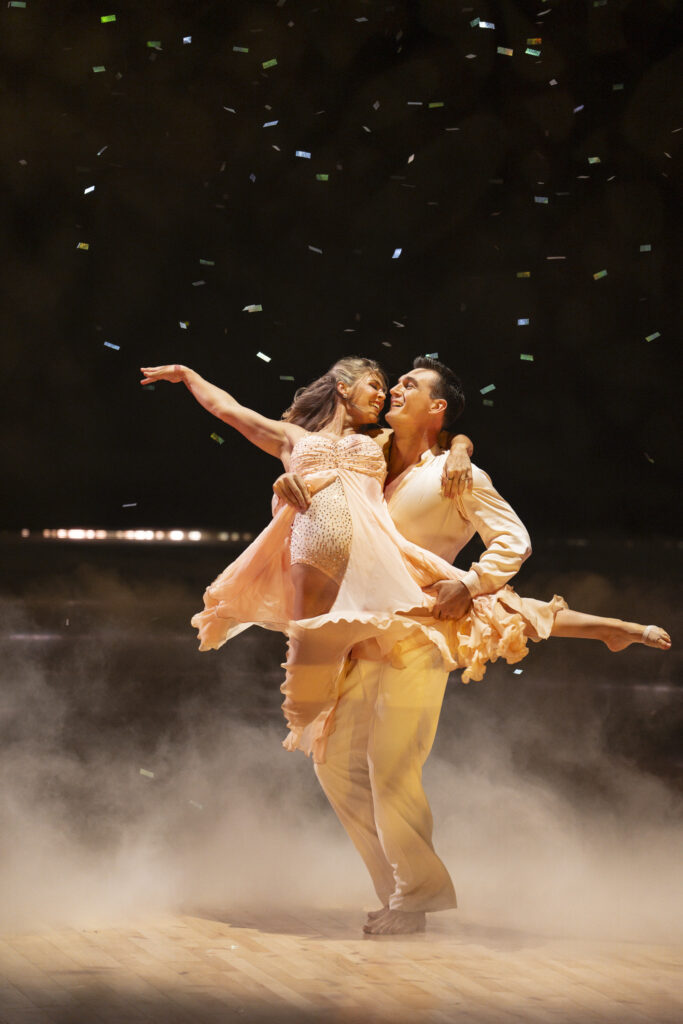 Onstage with mist on the floor and confetti fluttering down, Tyler Cameron lifts fiancée Tate Madden and spins her. Madden reaches an arm out while tucking one leg behind and extending the other out, foot pointed.
