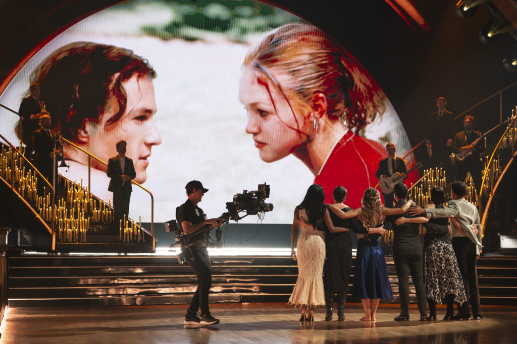 Onstage surrounded by candles and musicians, a group of "10 Things I Hate About You" cast members stand side-by-side, arms around each other, as they face away from the camera to look at an image projected at the back of the stage showing Julia Stiles and Heath Ledger in a still from the movie. A camera man stands next to them, recording.