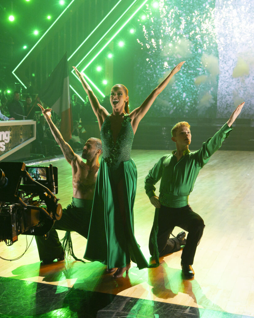 Mark Ballas, Maura Higgins, and Lochlan "Lucky" Howard pose triumphantly while bathed in green lighting, fireworks exploding behind them as a camera records in front. Higgins stands in the middle with her arms raised in a high "V" as the men kneel on one knee on either side, inside arms on hips and outside arms raised like Higgins'.