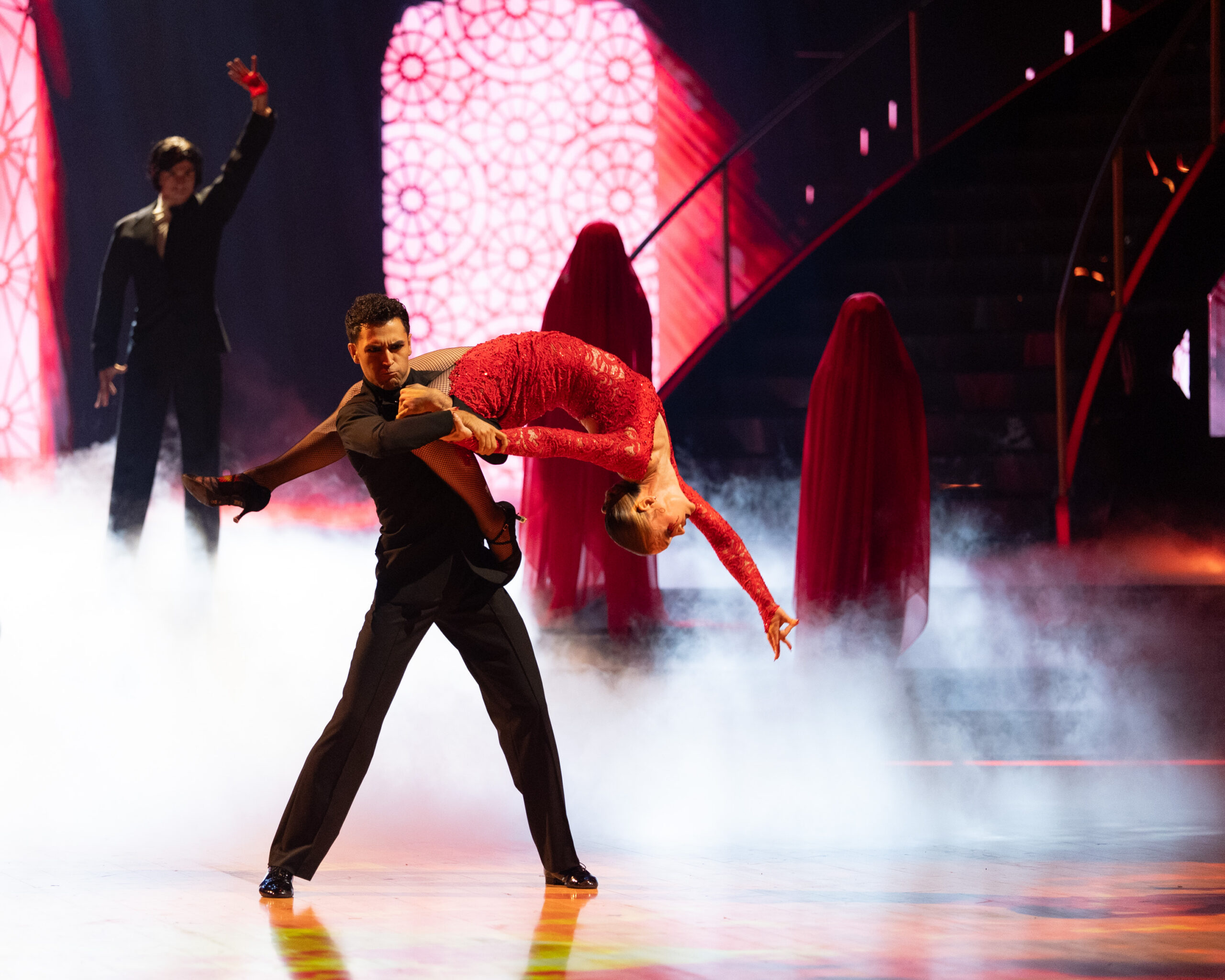 Ezra Sosa, in black, and Julia Stiles, in red, dance together on a misty stage with three figures in the back and stained-glass projections on the wall. Sosa stands wide-legged as he lifts Stiles, who arches backward with one leg behind him and her other wrapped around his arm. She reaches one arm back and toward the floor.