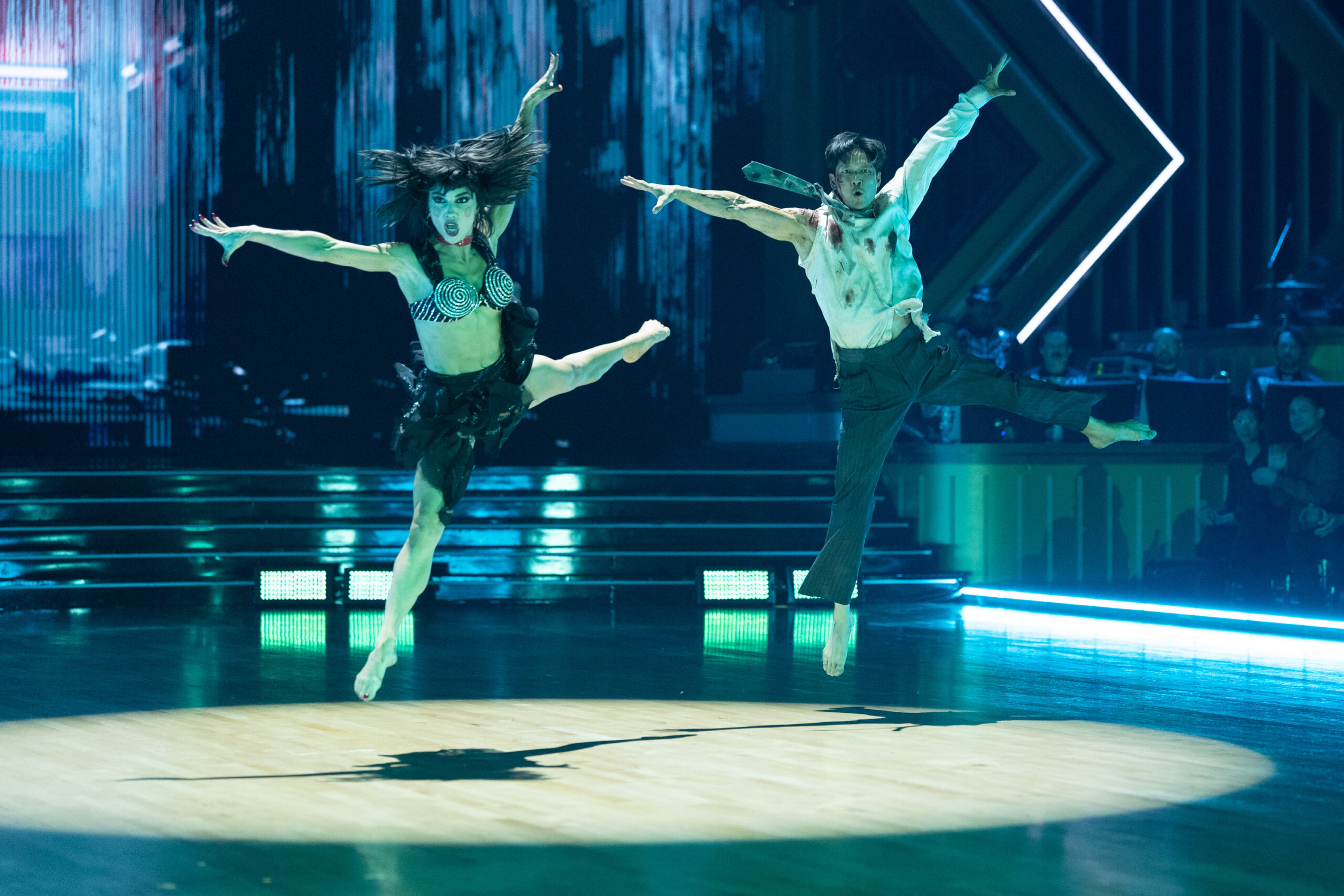 Jenna Johnson and Harry Shum Jr. sauté in open arabesques, arms wide, in a spotlight. Johnson wears a black skirt and bra top, and Shum Jr. wears dark pants and a bloody shirt and tie.