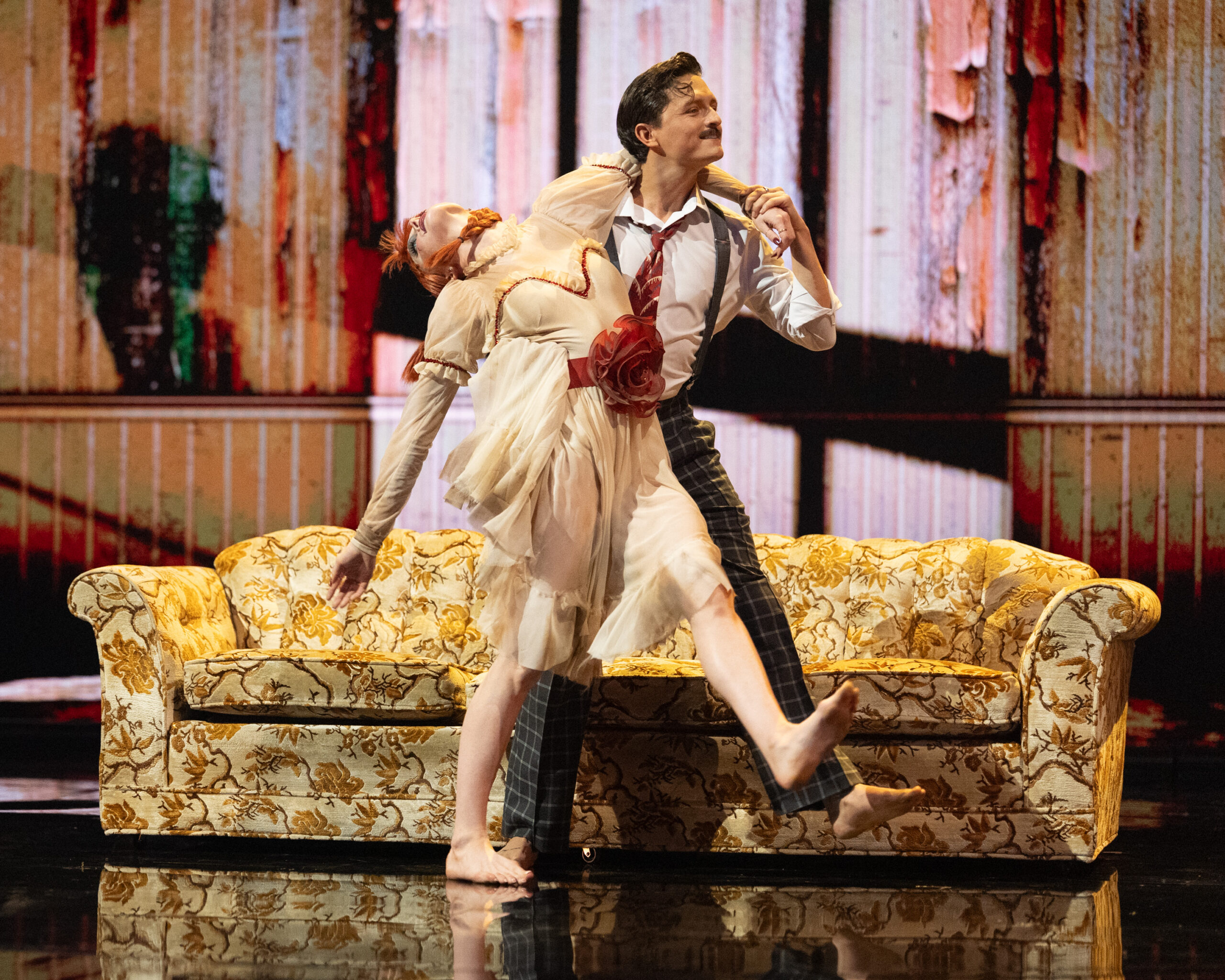 Connor Wood, in a dapper costume with suspenders, walks forward cheerily while supporting Rylee Arnold, who is dressed like a doll and flops back while stepping with Wood. They walk in front of a yellow couch.