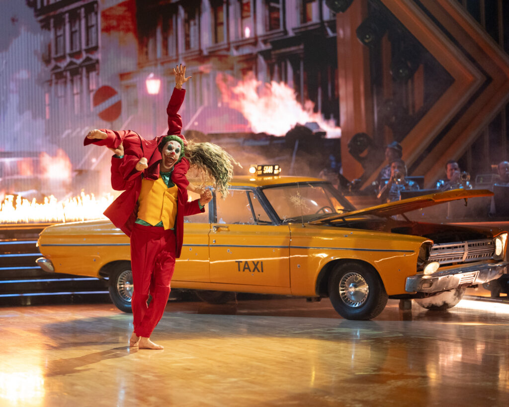 Jackson Olson and Emma Slater, dressed like the Joker with face paint, dance in front of a yellow taxi and a city backdrop with fires. Olson gives a freaky smile as he holds Slater across his shoulders with her back facing the camera.