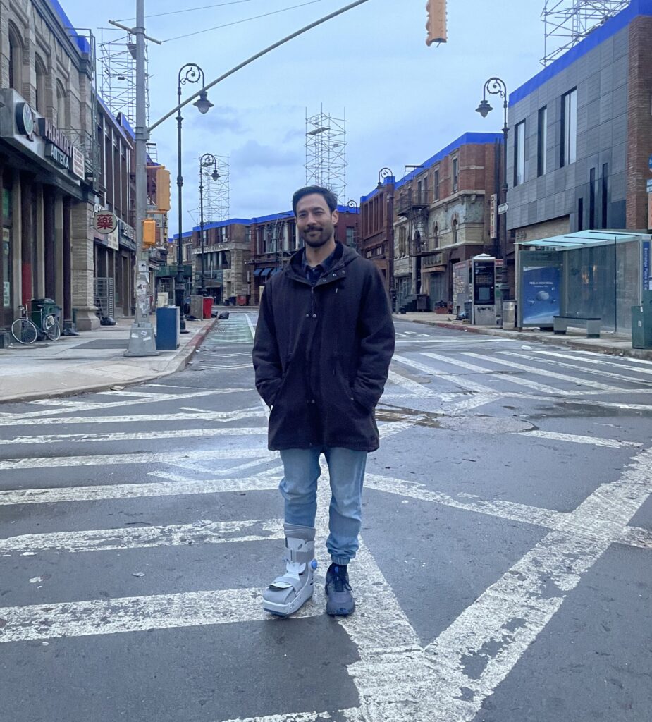 Nathan Kim stands on a crosswalk on a quiet street, hands in his coat pockets. He wears a medical boot on one foot.