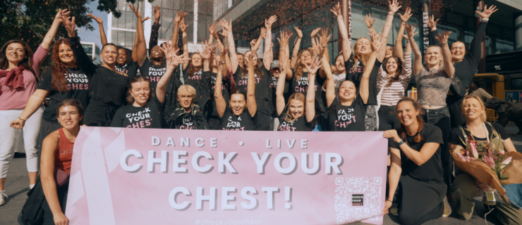 A large group of dancers celebrate and pose for a photo holding a large, light pink sign that reads "Dance, live, check your chest!"