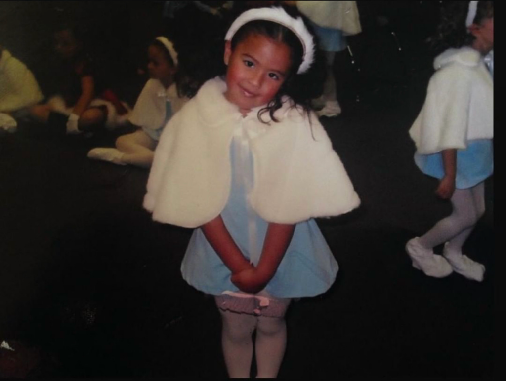 A childhood photo of Stephani Sosa backstage at a dance performance. She wears a powder blue dress with a fuzzy white shawl and a white headband. She stands with her legs together and her hands held in front, head slightly cocked as she gives a shy smile.