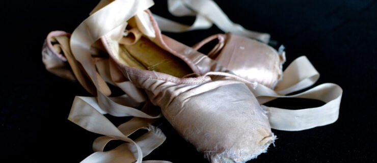 A dead pair of pointe shoes rest against a black background.