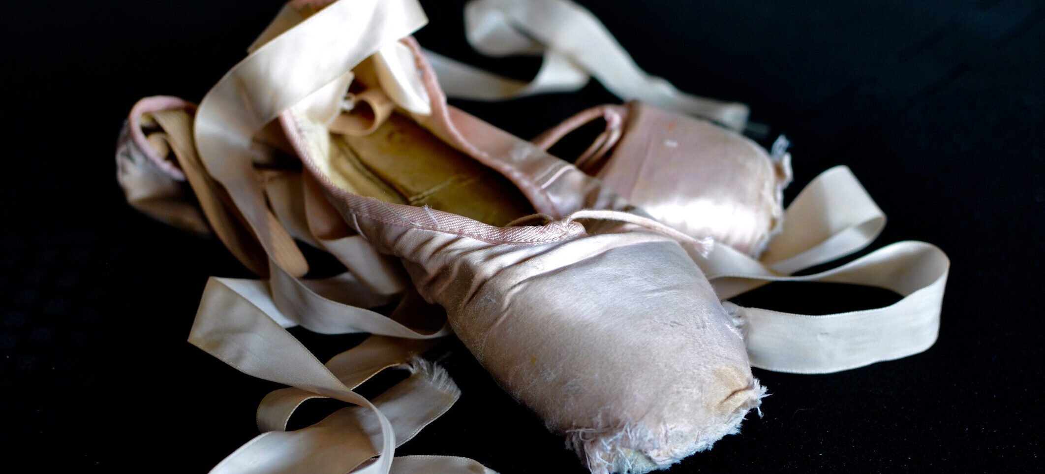 A dead pair of pointe shoes rest against a black background.