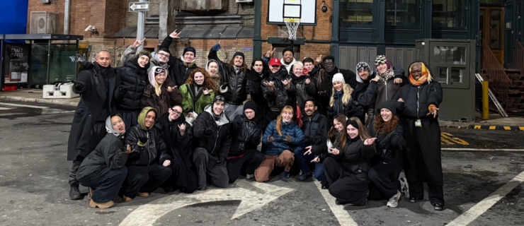 On the street at night, a large group of dancers pose together for a photo. They are bundled warmly with coats, hats, and scarves.