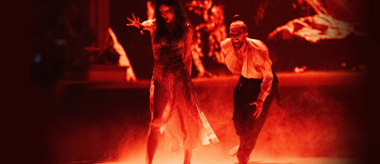 Maura Higgins and Mark Ballas dance in a spotlight, with the stage colored red. Higgins walks forward ominously, one arm extended front with flexed fingers, and her head tilted down with her mouth open. Ballas runs toward her from behind, mouth open to expose fangs.
