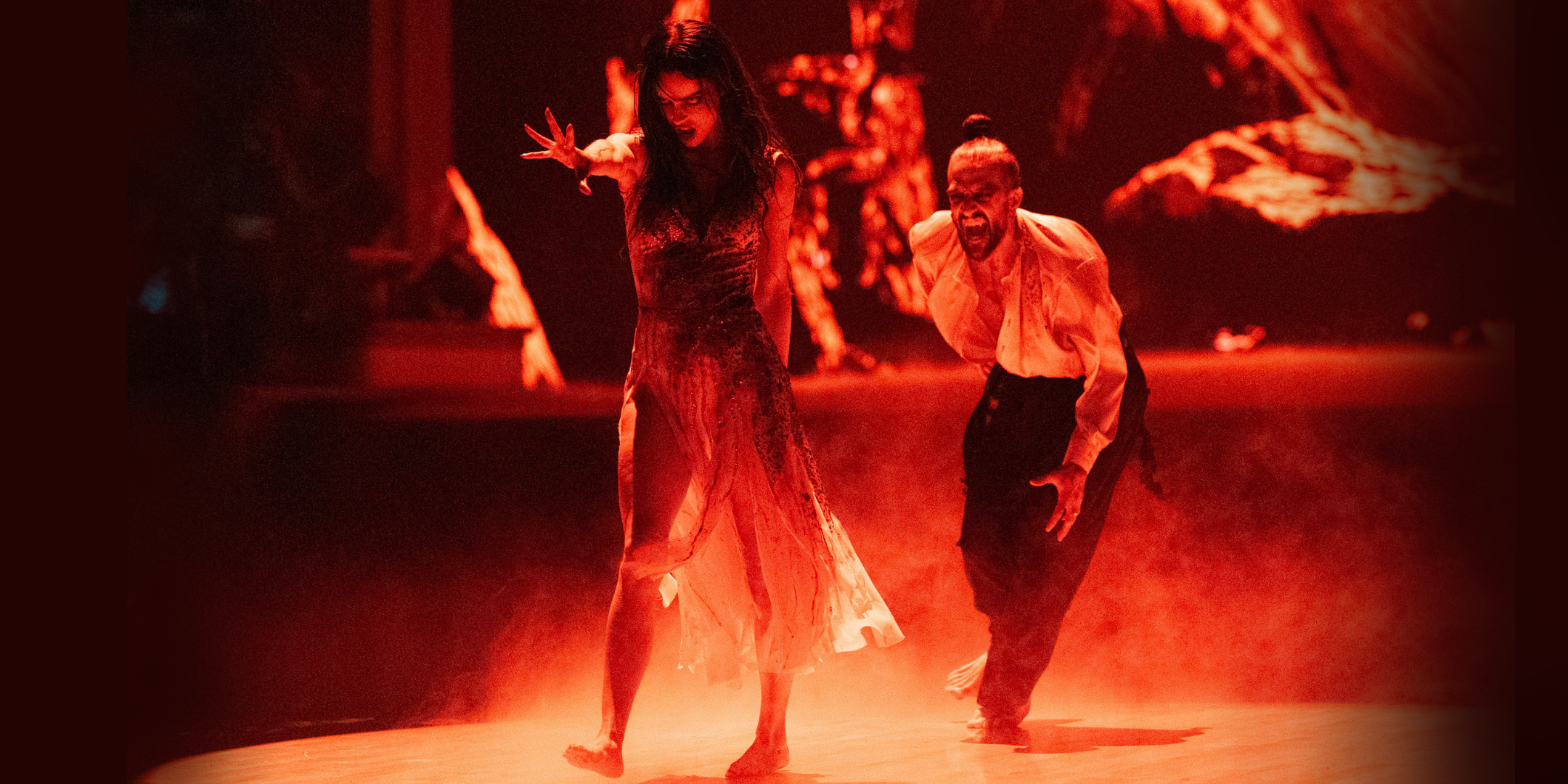 Maura Higgins and Mark Ballas dance in a spotlight, with the stage colored red. Higgins walks forward ominously, one arm extended front with flexed fingers, and her head tilted down with her mouth open. Ballas runs toward her from behind, mouth open to expose fangs.