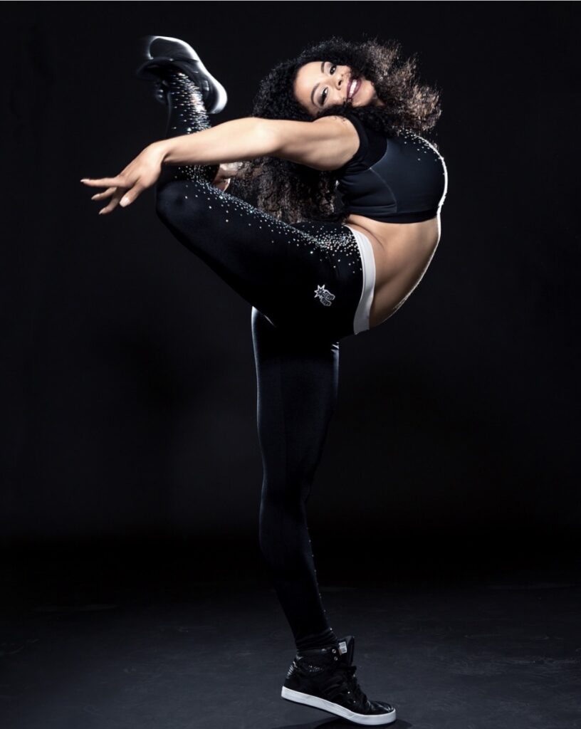 Jessica Butler wears a San Antonio Spurs dance team uniform and poses in front of a black backdrop. She kicks one leg back into a high attitude, arching back with her arms reaching behind her, while she smiles at the camera.
