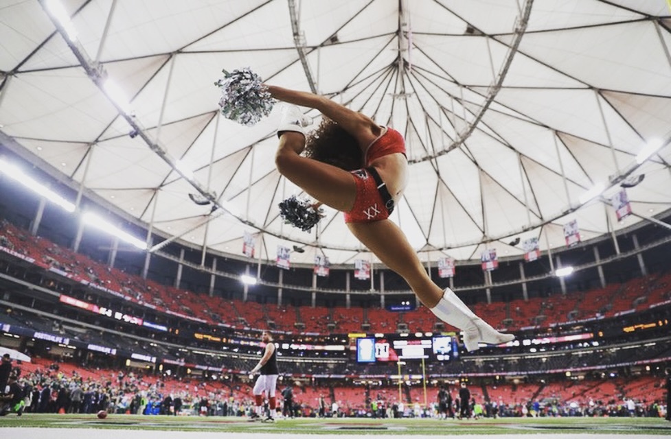 Jessica Butler wears a cheer uniform and does a Firebird jump in a massive arena. She holds silver poms in either hands.