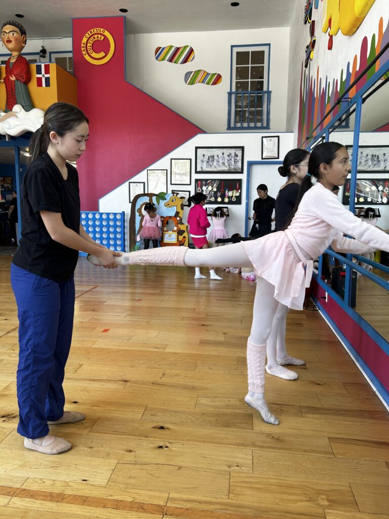 In a ballet studio with a pink wall, a teen dance instructor helps a young student with her arabesque at the barre.