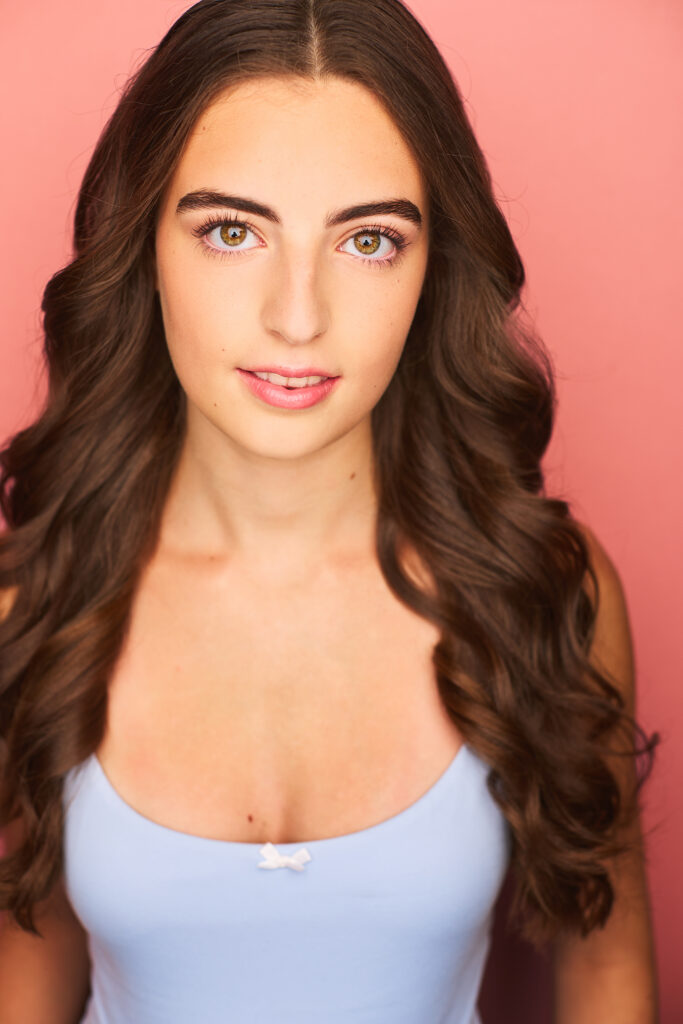 Isabella Addison Perrone's headshot. She wears a white shirt with her long hair down. The backdrop is pink.