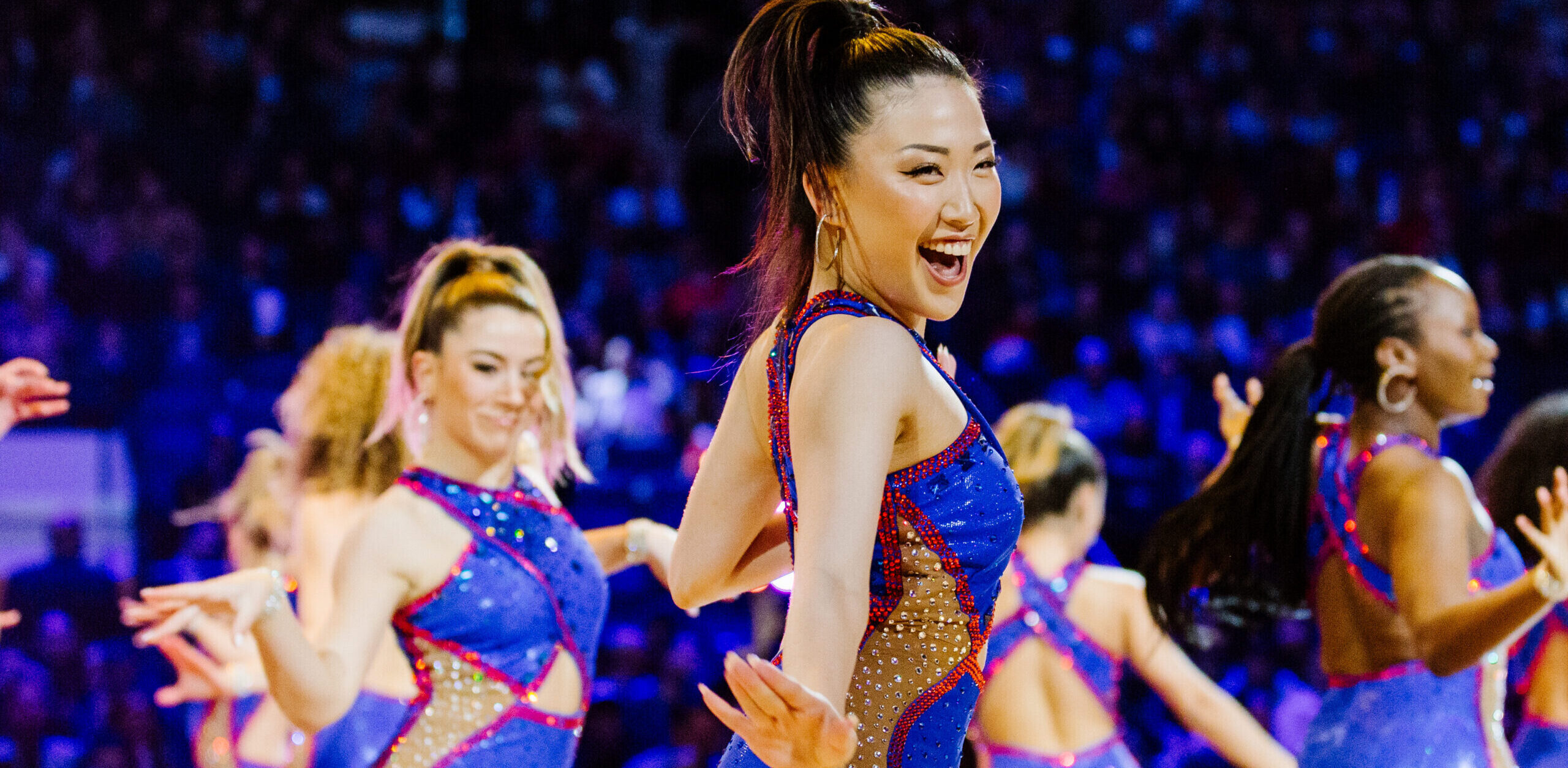 A dance team performs on a basketball court in tight, shiny costumes with a bright blue base and gold and red accents. They pop a hip, looking over their right shoulders, with one arm bent and the other extended out to the side, hands flexed.