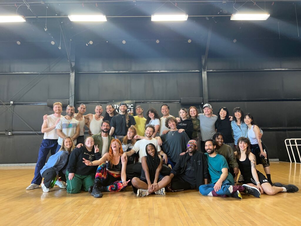 A large group of dancers pose together for a photo inside a large warehouse-style studio.