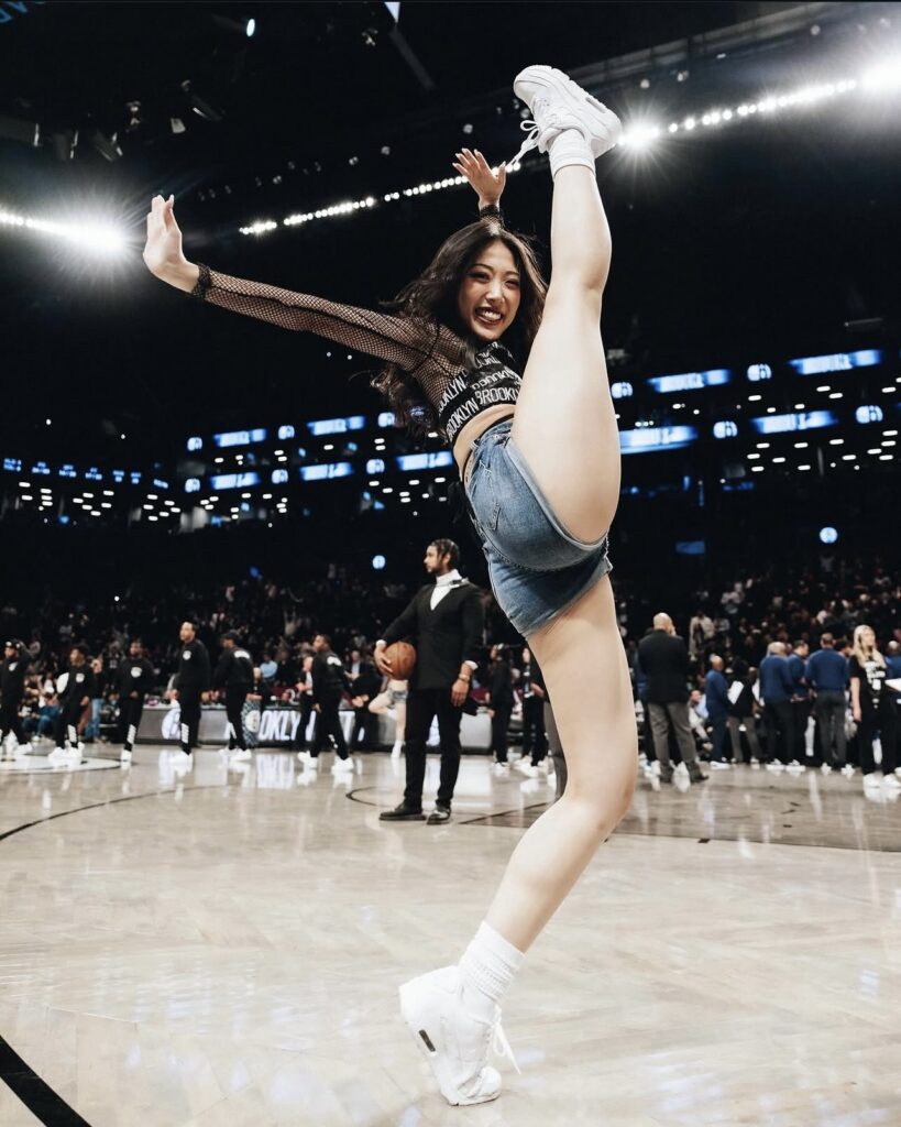 Hayoung Roh does a huge battement devant on a basketball court, smiling at the camera with one arm high and the other to the side. She wears a dark gray and black set with a mesh long-sleeved top, plus white tennis shoes.
