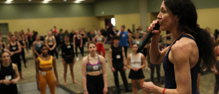 Suzi Taylor speaks into a microphone on a stage as she addresses a ballroom full of young dance students.