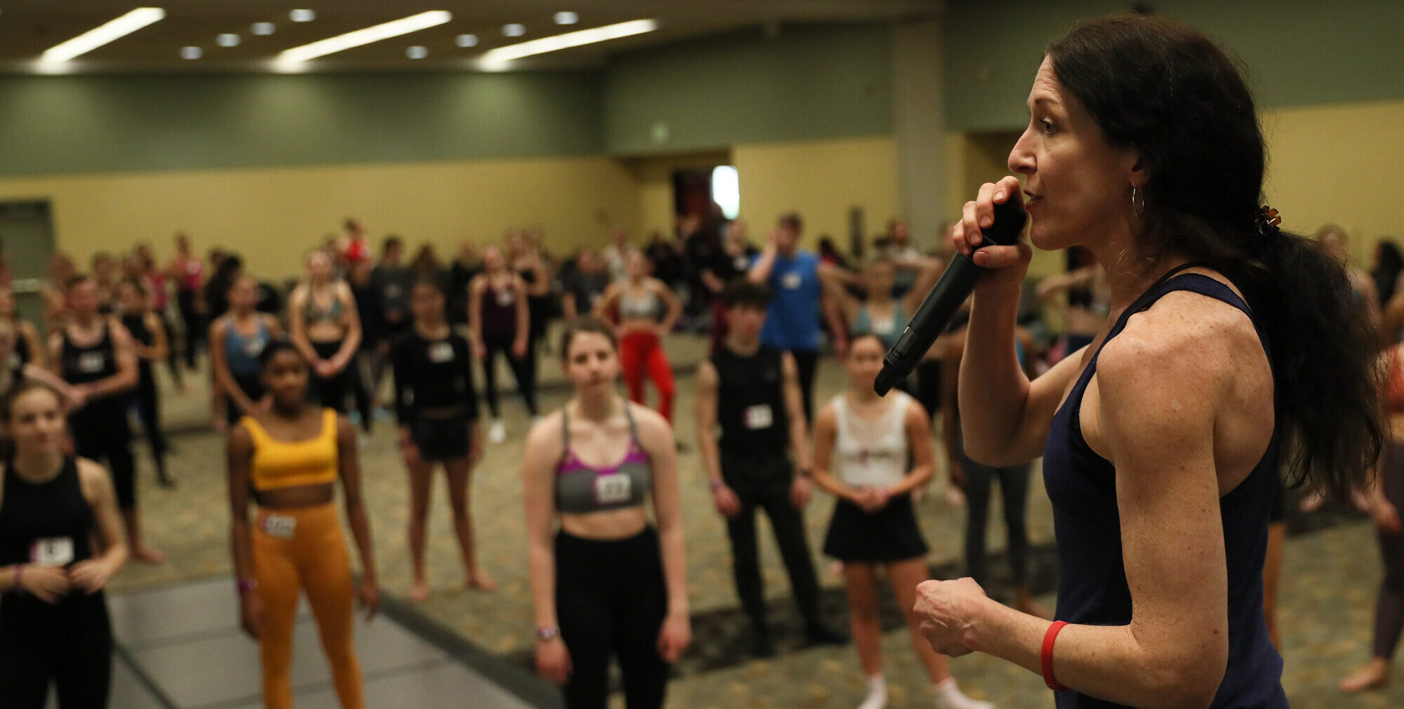 Suzi Taylor speaks into a microphone on a stage as she addresses a ballroom full of young dance students.