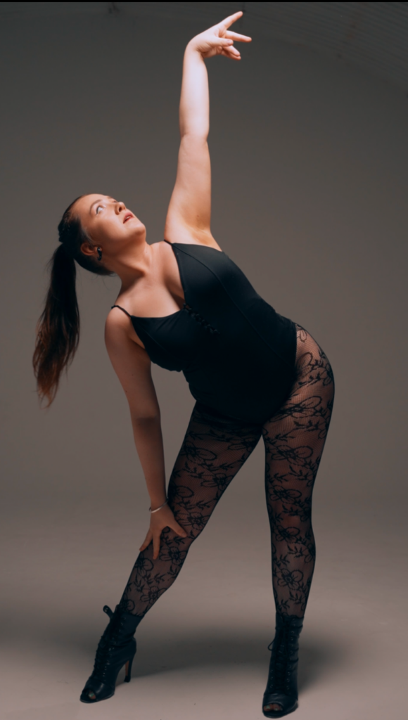 In front of a dark gray backdrop, Lucy Gray Freeman poses in a black leotard, black tights with floral patterns, and black heeled boots. She stands wide-legged and pops one hip, twisting her torso to reach up with one arm. She rests her other hand on her knee and looks up toward her top hand, her long hair in a high ponytail.