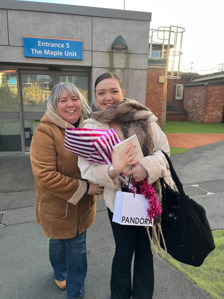 Lucy Mae Freeman poses with her mom outside a hospital. They are bundled in warm coats and lean in toward each other, smiling. Lucy Mae holds a large bag, a notebook, flowers, and a gift bag.